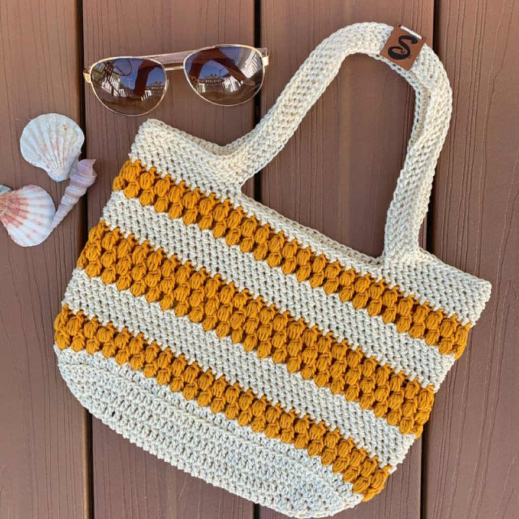 Ecru and gold crochet tote lies on a wooden deck. Sunglasses and seashells are close by.