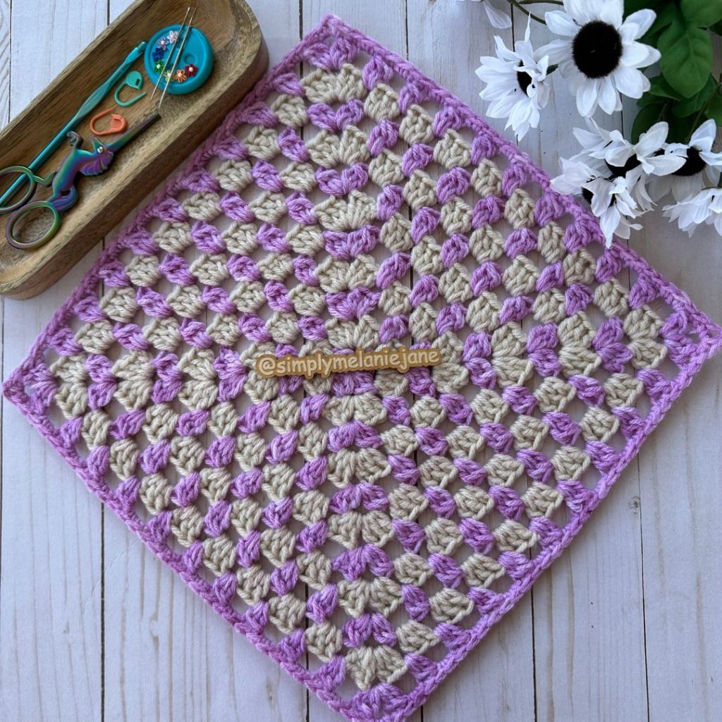 Purple and off white clustered granny square sits on a wooden background. A wooden container filled with crochet hook, scissors, stitch markers, and needles is nearby.